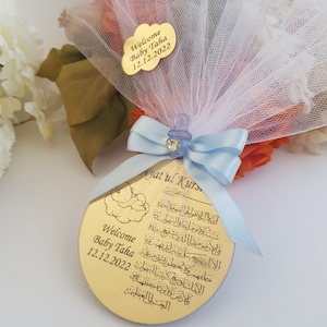 May include: A gold round ornament with Arabic calligraphy and the text "Welcome Baby Taha 12.12.2022" and "Ayatul Kurs"  wrapped in a blue ribbon and white tulle.