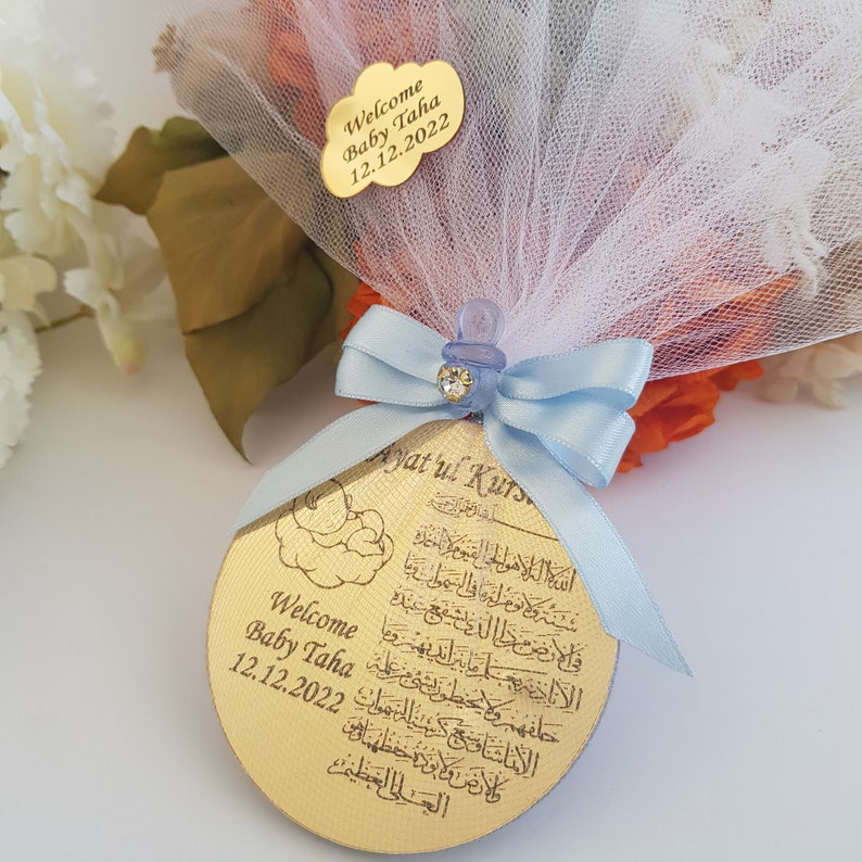 May include: A gold round ornament with Arabic calligraphy and the text "Welcome Baby Taha 12.12.2022" and "Ayatul Kurs"  wrapped in a blue ribbon and white tulle.