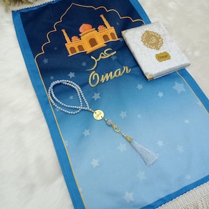 May include: A blue prayer mat featuring a mosque illustration, the name Omar in Arabic and English, and white star patterns. It includes a white textured book with a gold emblem and the name Omar, plus a pearl prayer bead strand with a gold charm.