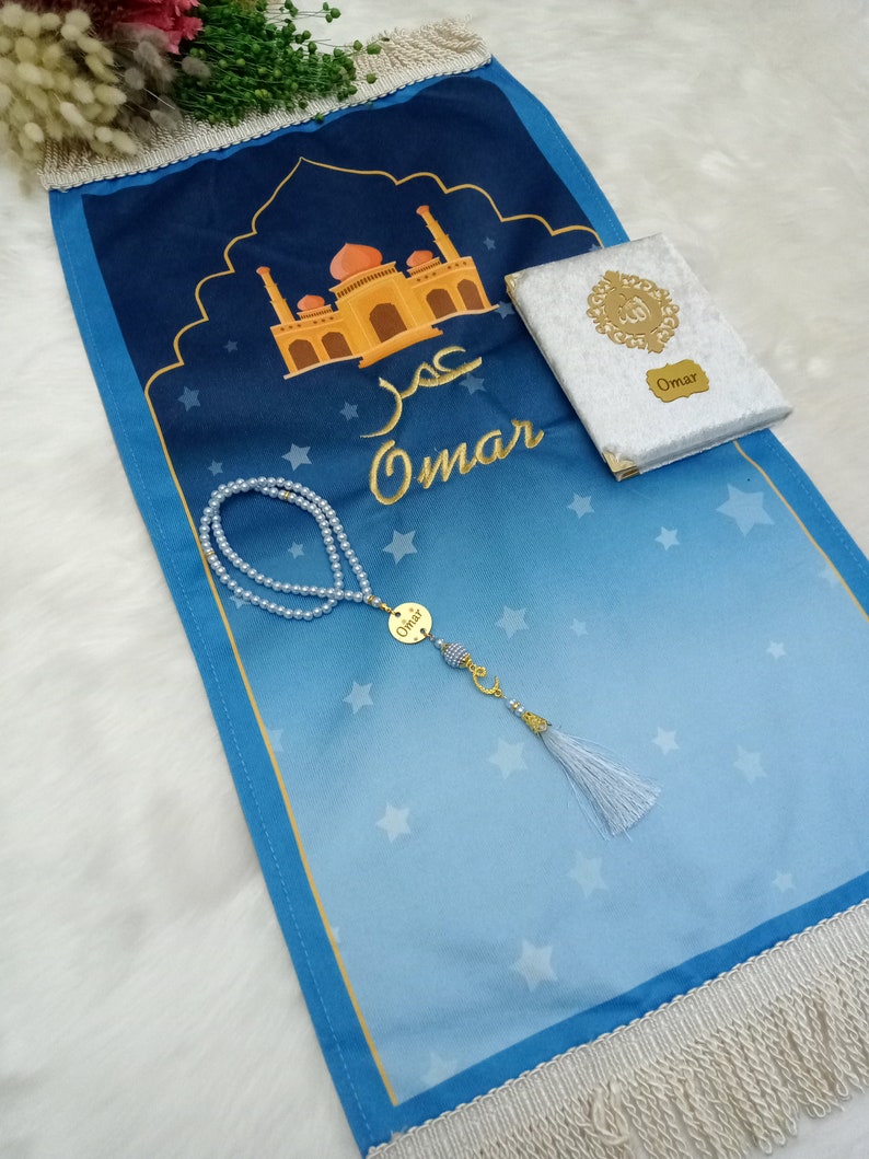 May include: A blue prayer mat featuring a mosque illustration, the name Omar in Arabic and English, and white star patterns. It includes a white textured book with a gold emblem and the name Omar, plus a pearl prayer bead strand with a gold charm.