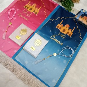May include: Two prayer mats with fringe, one pink and one blue, featuring mosque illustrations and the names Lyanna and Omar. Each set includes a matching prayer bead strand and a small book with a gold-toned emblem and name tag.
