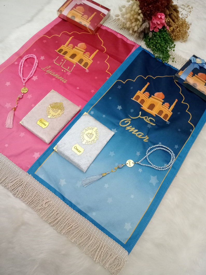 May include: Two prayer mats with fringe, one pink and one blue, featuring mosque illustrations and the names Lyanna and Omar. Each set includes a matching prayer bead strand and a small book with a gold-toned emblem and name tag.
