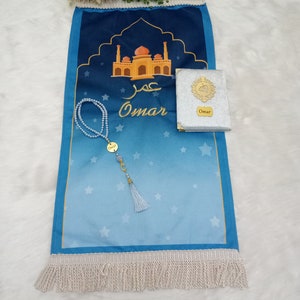 May include: A blue prayer mat featuring a mosque illustration, the name Omar in Arabic and English script, and white fringe. A set of white pearl prayer beads with a gold Omar charm and a small silver book with a gold emblem and Omar label.