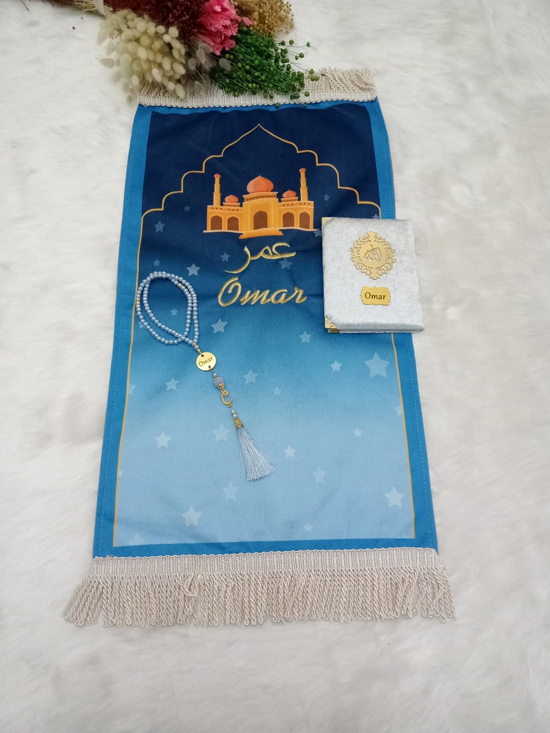 May include: A blue prayer mat featuring a mosque illustration, the name Omar in Arabic and English script, and white fringe. A set of white pearl prayer beads with a gold Omar charm and a small silver book with a gold emblem and Omar label.