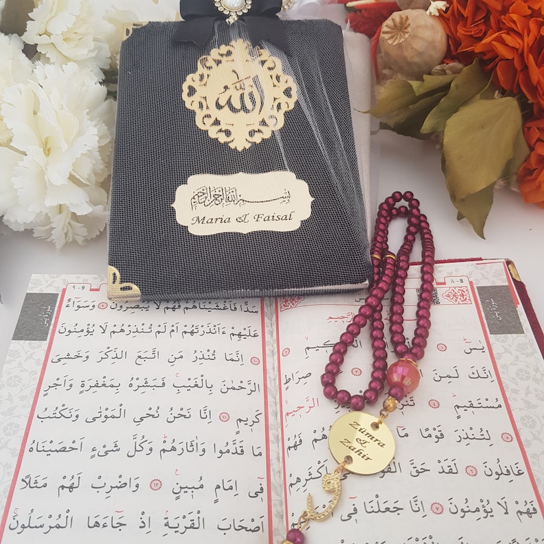 Pode incluir: Um livro coberto de tecido preto com detalhes dourados e escrita &aacute;rabe, personalizado com os nomes "Maria el Faisal". Uma corrente de ora&ccedil;&atilde;o com contas bord&ocirc; e um Alcor&atilde;o aberto tamb&eacute;m s&atilde;o vis&iacute;veis. Um pingente dourado com escrita &aacute;rabe pende da corrente.