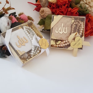 May include: Two small gift boxes with gold trim and clear lids. Each box contains a white fabric square with gold Arabic calligraphy, a string of white or brown beads, and a small gold tag. One tag reads "Abdullah & Razan" and the other reads "Mohammad & Khalifah 5.14.2022".