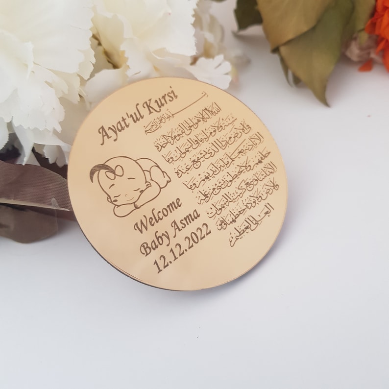 May include: A round, wooden plaque with a gold finish. The plaque features a sleeping baby illustration, the text "Ayat'ul Kursi" in Arabic script, and the words "Welcome Baby Asma 12.12.2022".