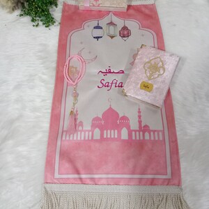 May include: A pink prayer rug with a white fringe, a pink velvet prayer book with gold trim, a pink prayer bead necklace, and a small gift box with a pink ribbon. The prayer rug has a design of a mosque with minarets and lanterns. The prayer book has the name "Safia" on the cover. The gift box has the word "Nora" on the top.