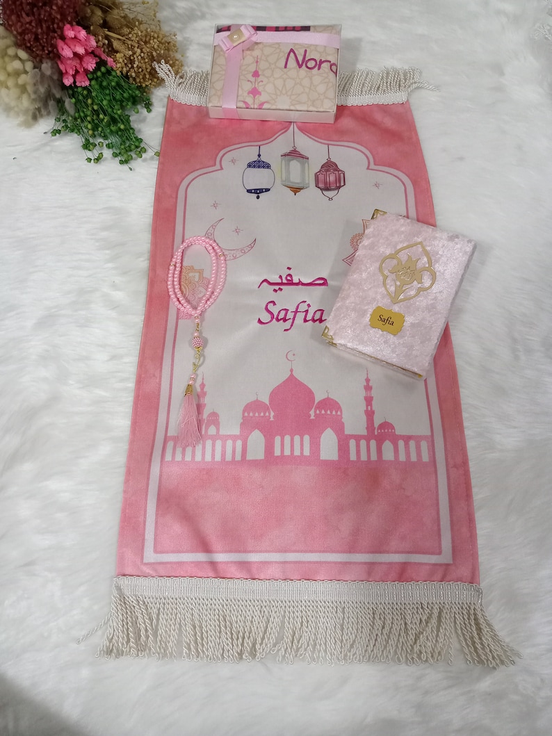May include: A pink prayer rug with a white fringe, a pink velvet prayer book with gold trim, a pink prayer bead necklace, and a small gift box with a pink ribbon. The prayer rug has a design of a mosque with minarets and lanterns. The prayer book has the name "Safia" on the cover. The gift box has the word "Nora" on the top.