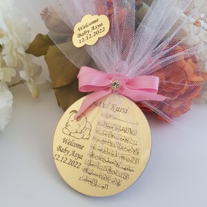 May include: A gold-colored round wooden ornament with a baby elephant design and Arabic calligraphy. The ornament is decorated with a pink ribbon and a clear plastic charm. The text on the ornament reads "Welcome Baby Asya 12.12.2022" and "Ayatul Kursi".