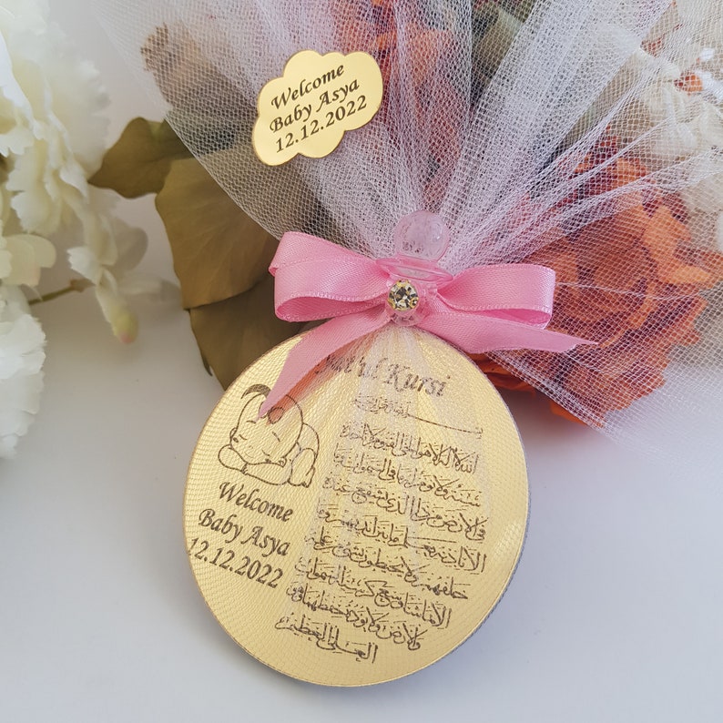 May include: A gold-colored round wooden ornament with a baby elephant design and Arabic calligraphy. The ornament is decorated with a pink ribbon and a clear plastic charm. The text on the ornament reads "Welcome Baby Asya 12.12.2022" and "Ayatul Kursi".