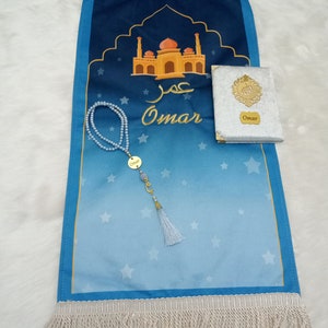May include: A blue prayer mat with a gradient design, white stars, and a gold mosque illustration. It features the name Omar in Arabic and English script. Included are a white velvet-covered book with a gold emblem and a pearl prayer bead strand.