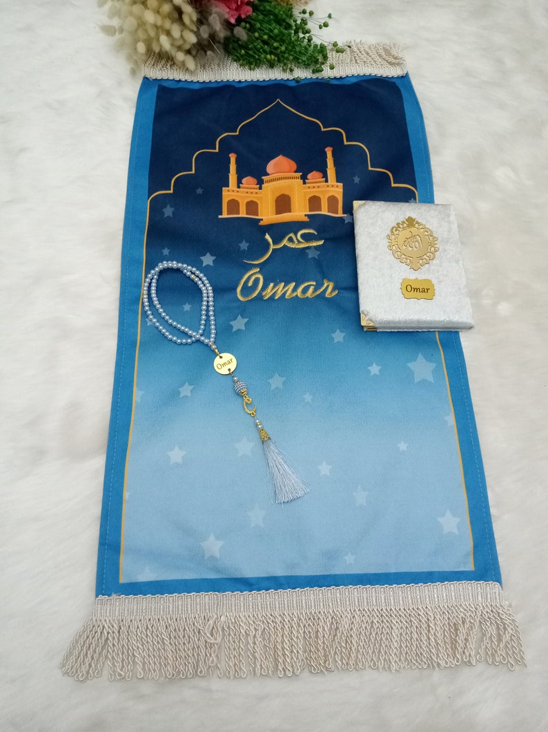 May include: A blue prayer mat with a gradient design, white stars, and a gold mosque illustration. It features the name Omar in Arabic and English script. Included are a white velvet-covered book with a gold emblem and a pearl prayer bead strand.