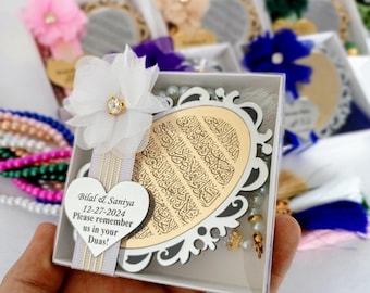 May include: A white gift box with a gold trim and a clear plastic window. Inside the box is a white wooden oval with gold Arabic calligraphy. The oval is attached to a white heart-shaped plaque with the text "Bilal & Saniya 12-27-2024 Please remember us in your Duas!" The box also contains a white string of beads and a white tassel.