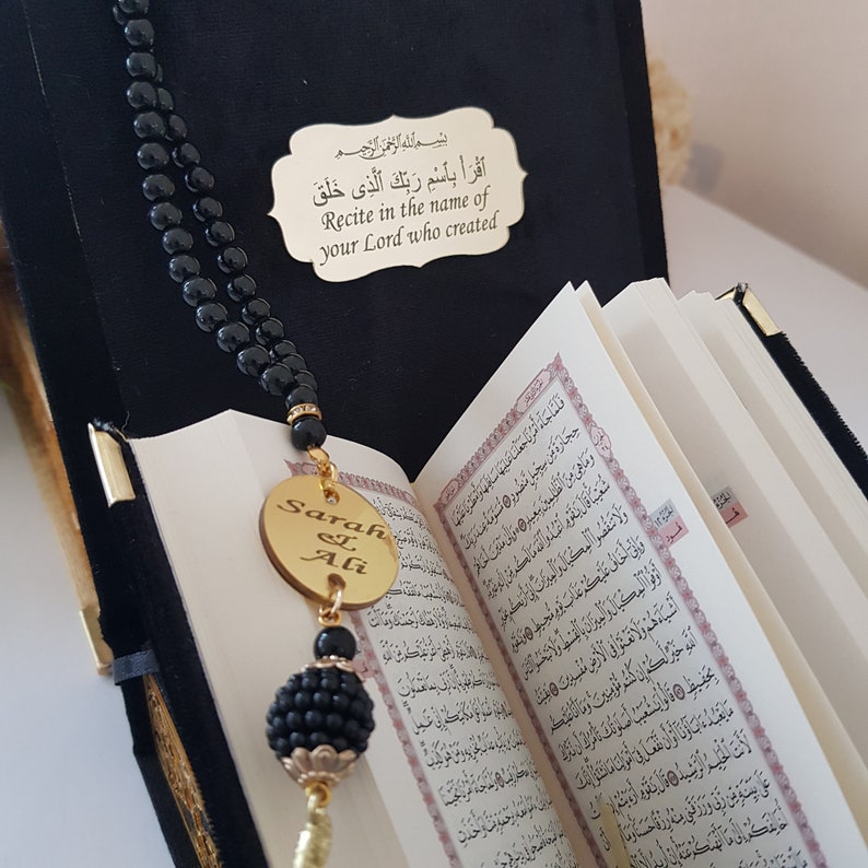 May include: A black prayer beads necklace with a gold pendant engraved with the names "Sarah & Ali". The pendant is hanging over an open Quran with Arabic text. The text on the plaque reads "Recite in the name of your Lord who created".