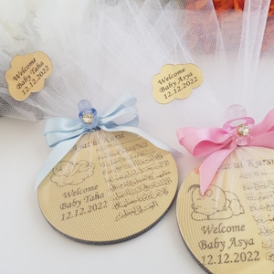 May include: Two round wooden ornaments with a baby design and Arabic text. The ornaments are decorated with blue and pink ribbons and pacifiers. The text on the ornaments reads "Welcome Baby Taha 12.12.2022" and "Welcome Baby Asya 12.12.2022".