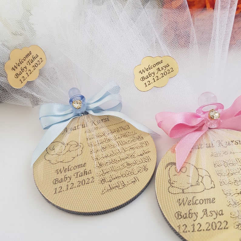 May include: Two round wooden ornaments with a baby design and Arabic text. The ornaments are decorated with blue and pink ribbons and pacifiers. The text on the ornaments reads "Welcome Baby Taha 12.12.2022" and "Welcome Baby Asya 12.12.2022".