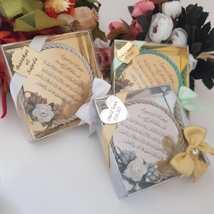 May include: Three wedding favours in clear boxes. Each features a circular design with Arabic script, a rope border, and floral decorations. The boxes are tied with ribbons and have personalised tags with names and dates.