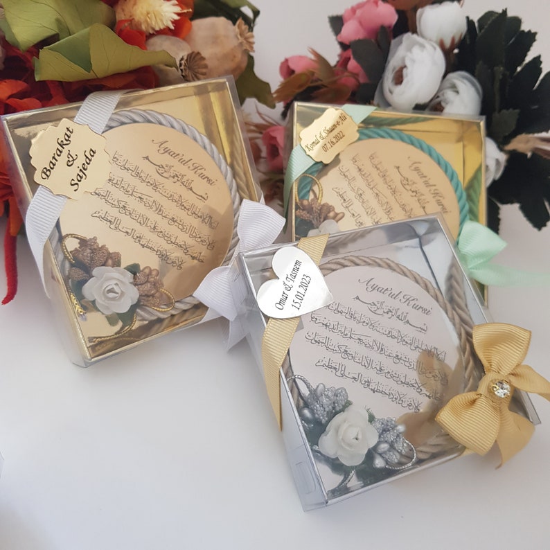 May include: Three wedding favours in clear boxes. Each features a circular design with Arabic script, a rope border, and floral decorations. The boxes are tied with ribbons and have personalised tags with names and dates.