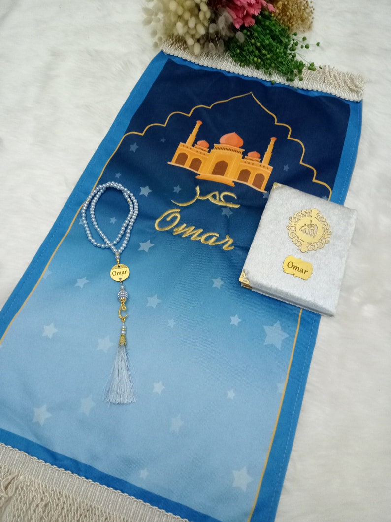 May include: A blue prayer mat with a white star pattern, a mosque illustration, and the name Omar in gold embroidery. It includes a set of light blue pearl prayer beads with a gold crescent charm and a silver-colored book with a gold name tag.