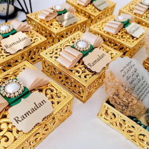 Op de afbeelding: Gouden decoratieve dozen met ingewikkelde ontwerpen, voorzien van een groene en witte strik met een parelversiering. Elke doos heeft een label met de tekst "Ramadan".