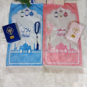 May include: Two blue and pink prayer rugs with white fringe, each with a mosque design, a crescent moon, and a string of beads. The rugs are labeled "Bilal" and "Safia".