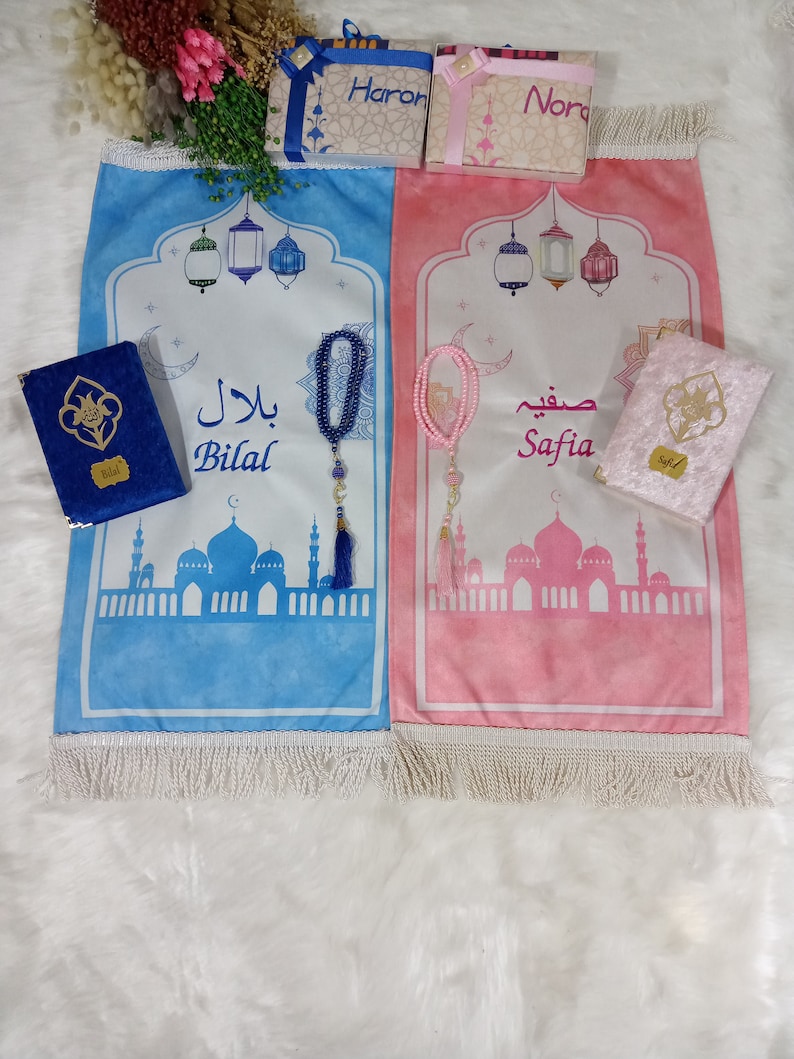 May include: Two blue and pink prayer rugs with white fringe, each with a mosque design, a crescent moon, and a string of beads. The rugs are labeled "Bilal" and "Safia".
