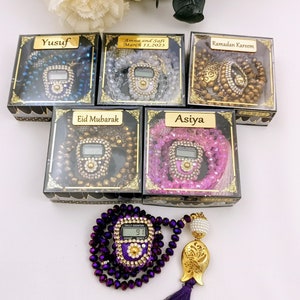 May include: Five small gift boxes each containing a digital prayer bead counter with a beaded necklace. The boxes are labeled with names and dates. The prayer bead counters are in different colors, including blue, white, brown, gold, and pink.