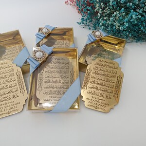 Pu&ograve; includere: Quattro scatole regalo dorate con nastro blu e decorazioni di perle. Ogni scatola contiene una placca dorata con calligrafia araba e il testo "Al-Fatiha" e "Benvenuto Baby Shahzain 09.13.2022".