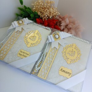 May include: Two gift boxes with white books, gold accents, and pearl rosaries. Each box is tied with a white ribbon and features a gold emblem and a personalized name tag. Dried flowers in various colors are in the background.