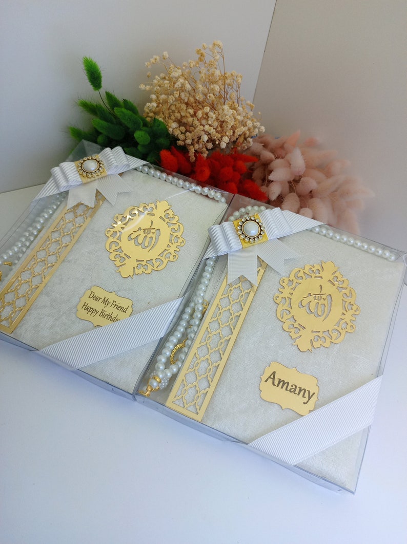 May include: Two gift boxes with white books, gold accents, and pearl rosaries. Each box is tied with a white ribbon and features a gold emblem and a personalized name tag. Dried flowers in various colors are in the background.