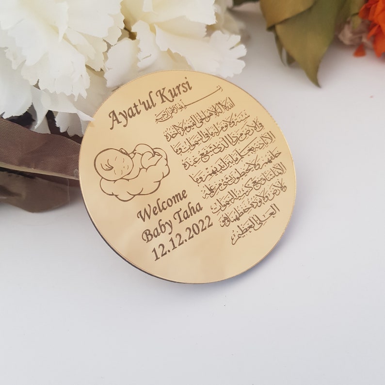May include: A gold-colored round plaque with an engraved image of a baby sleeping on a cloud. The plaque also features the Arabic text of the Ayat'ul Kursi, a verse from the Quran, and the text "Welcome Baby Taha 12.12.2022".