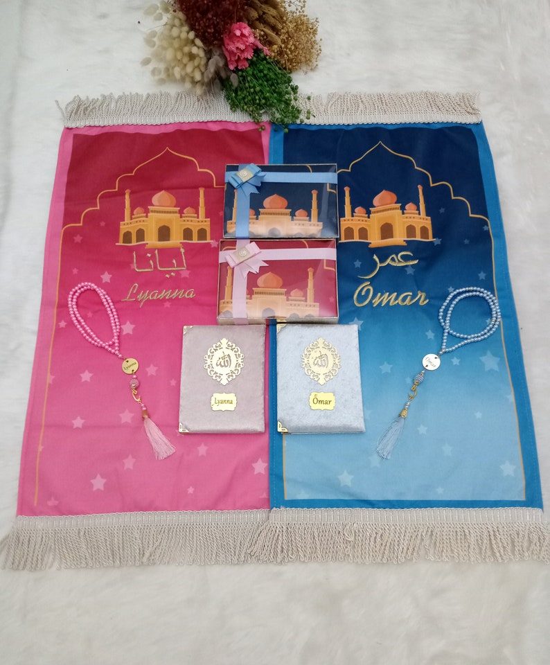 May include: Two prayer mats in pink and blue featuring mosque illustrations and the names Lyanna and Omar. Includes two matching gift boxes with ribbons, two velvet-covered books with gold calligraphy, and two sets of beaded prayer beads with tassels.