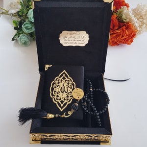 May include: A black velvet box with gold trim, containing a small black prayer book with gold Arabic calligraphy, a black prayer bead necklace, and a gold charm with the word "Sarah" engraved on it. The box lid has a gold plaque with the Arabic text "أَقْرَأْ بِاسْمِ رَبِّكَ الَّذِي خَلَقَ" and the English translation "Recite in the name of your Lord who created."