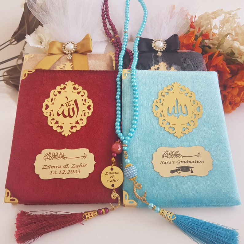Pode incluir: Dois livros de veludo, um vermelho e outro azul, com detalhes dourados e caligrafia &aacute;rabe. Cada livro tem um elemento decorativo dourado e uma placa dourada personalizada. Um colar de contas e uma borla est&atilde;o sobre os livros.