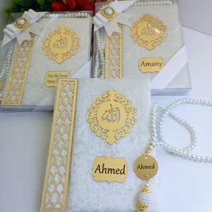 May include: White velvet-covered books with gold embellishments, including a decorative emblem and nameplates. Each book is packaged in a clear box and adorned with a white ribbon and pearl-like beads. The text "Ahmed" is visible.