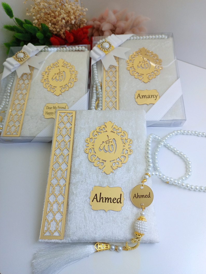 May include: White velvet-covered books with gold embellishments, including a decorative emblem and nameplates. Each book is packaged in a clear box and adorned with a white ribbon and pearl-like beads. The text "Ahmed" is visible.