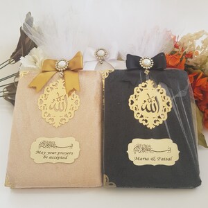 Pode incluir: Dois livros decorativos, um bege e outro preto, s&atilde;o adornados com detalhes dourados e la&ccedil;os elegantes. Cada livro apresenta um recorte dourado com caligrafia &aacute;rabe e uma placa dourada com texto. O livro bege diz "May your prayers be accepted."