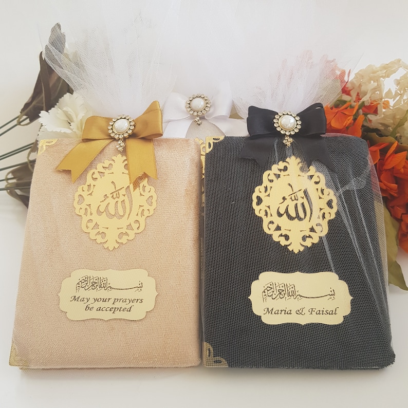 Pode incluir: Dois livros decorativos, um bege e outro preto, s&atilde;o adornados com detalhes dourados e la&ccedil;os elegantes. Cada livro apresenta um recorte dourado com caligrafia &aacute;rabe e uma placa dourada com texto. O livro bege diz "May your prayers be accepted."