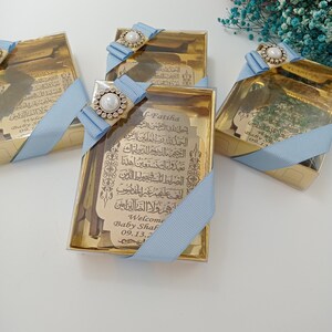 Pu&ograve; includere: Tre scatole dorate con nastro blu e decorazioni di perle. Ogni scatola contiene una piccola placca di legno con calligrafia araba e il testo "Welcome Baby Shaf 09.13.2".