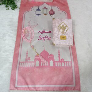 May include: A pink prayer rug with a white border and fringe. The rug features a mosque silhouette, lanterns, a crescent moon, and the name "Safia" in Arabic script. A pink velvet prayer book with the name "Safia" on the cover is included, along with a pink prayer beads.