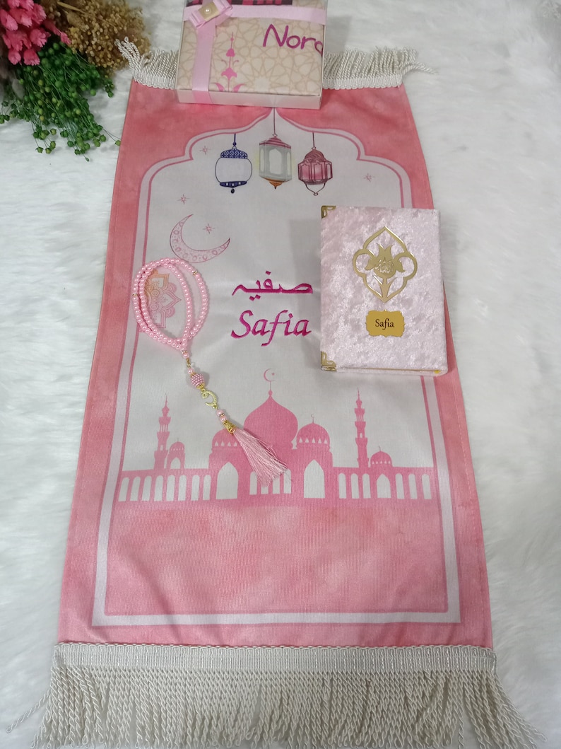 May include: A pink prayer rug with a white border and fringe. The rug features a mosque silhouette, lanterns, a crescent moon, and the name "Safia" in Arabic script. A pink velvet prayer book with the name "Safia" on the cover is included, along with a pink prayer beads.