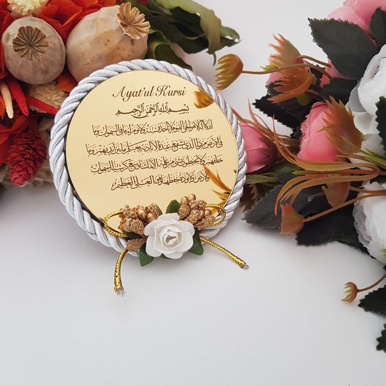 May include: A circular gold-coloured plaque featuring Arabic calligraphy, framed by a silver and white rope trim. A small floral arrangement with white and gold details is at the base. The background includes pink and white flowers.