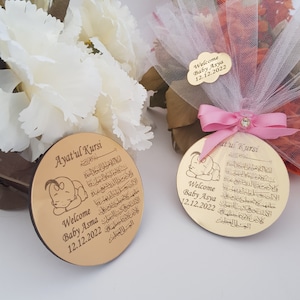 May include: Two round wooden plaques with a gold finish. Each plaque features a small illustration of a sleeping baby and Arabic text. The text on the plaques reads "Ayatul Kursi". The plaques also include the text "Welcome Baby Asma 12.12.2022" and "Welcome Baby Asya 12.12.2022".
