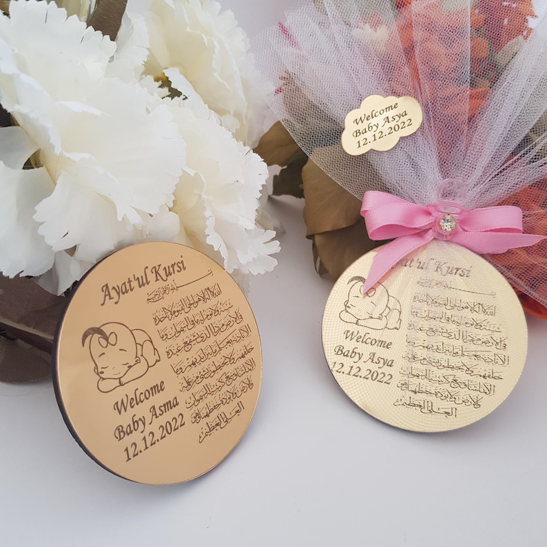 May include: Two round wooden plaques with a gold finish. Each plaque features a small illustration of a sleeping baby and Arabic text. The text on the plaques reads "Ayatul Kursi". The plaques also include the text "Welcome Baby Asma 12.12.2022" and "Welcome Baby Asya 12.12.2022".