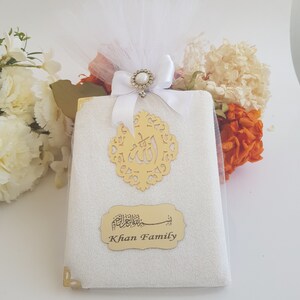 Pode incluir: Um presente embrulhado com um tecido branco, enfeites dourados e um la&ccedil;o branco. O presente &eacute; adornado com um recorte dourado com caligrafia &aacute;rabe e um r&oacute;tulo que diz "Khan Family". O fundo inclui flores brancas e laranjas.