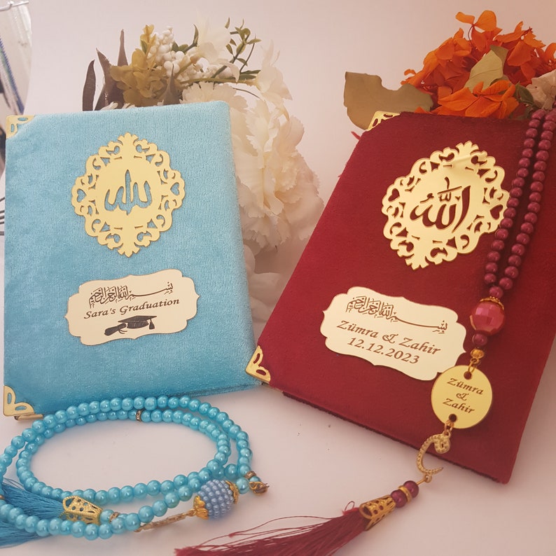 Pode incluir: Dois livros de veludo, um azul claro e outro bord&ocirc;, com detalhes dourados e caligrafia &aacute;rabe. O livro azul claro tem "Sara's Graduation" em uma placa dourada. Um ros&aacute;rio de contas azul claro est&aacute; em primeiro plano. O livro bord&ocirc; tem um ros&aacute;rio.