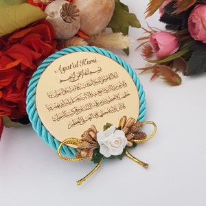 May include: A circular, gold-toned decorative item with Arabic script, encircled by a turquoise rope. It has a floral arrangement with a white rose and gold details. The text reads "Ayat'ul Kursi".