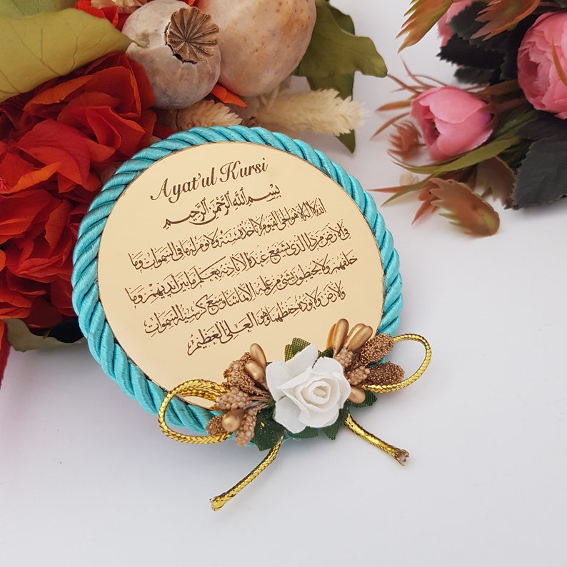 May include: A circular, gold-toned decorative item with Arabic script, encircled by a turquoise rope. It has a floral arrangement with a white rose and gold details. The text reads "Ayat'ul Kursi".