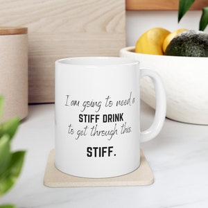 May include: White ceramic coffee mug with black text that reads "I am going to need a STIFF DRINK to get through this. STIFF."